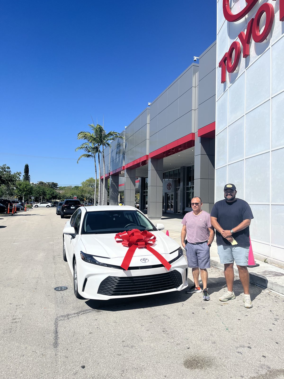 Two customers standing beside a white sedan with a red bow outside a Toyota dealership.
