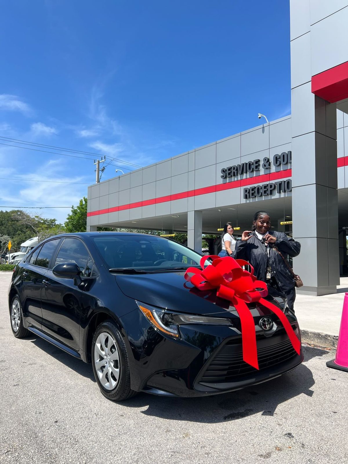 Customer picking up a black sedan with a red bow outside a dealership service center.